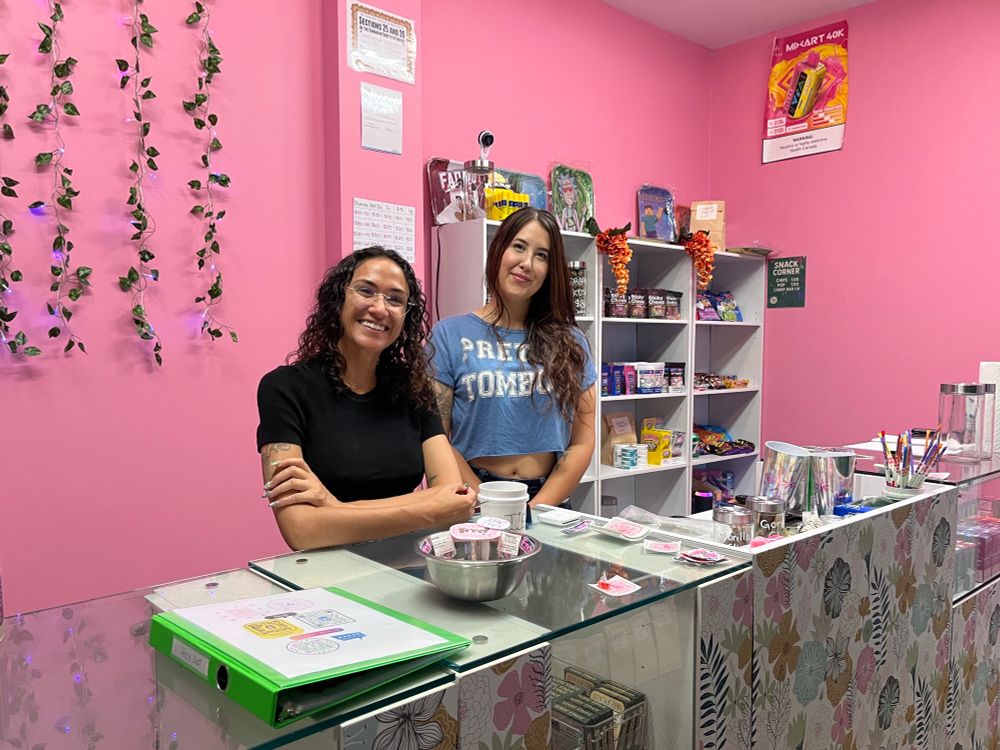Two people behind a retail counter with a pink wall behind them decorated with vines and shelves of product.