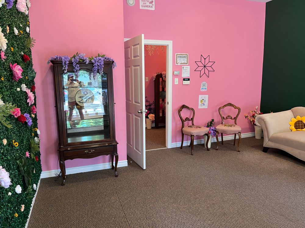 A room containing a sofa, two chairs, and a cabinet. The back wall is pink, side wall green, and end wall covered in artificial vines, leaves and flowers.