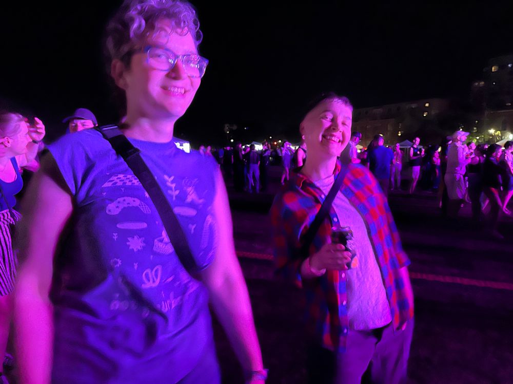 Two people pose in an ourdoor event space bathed in pink stage light.