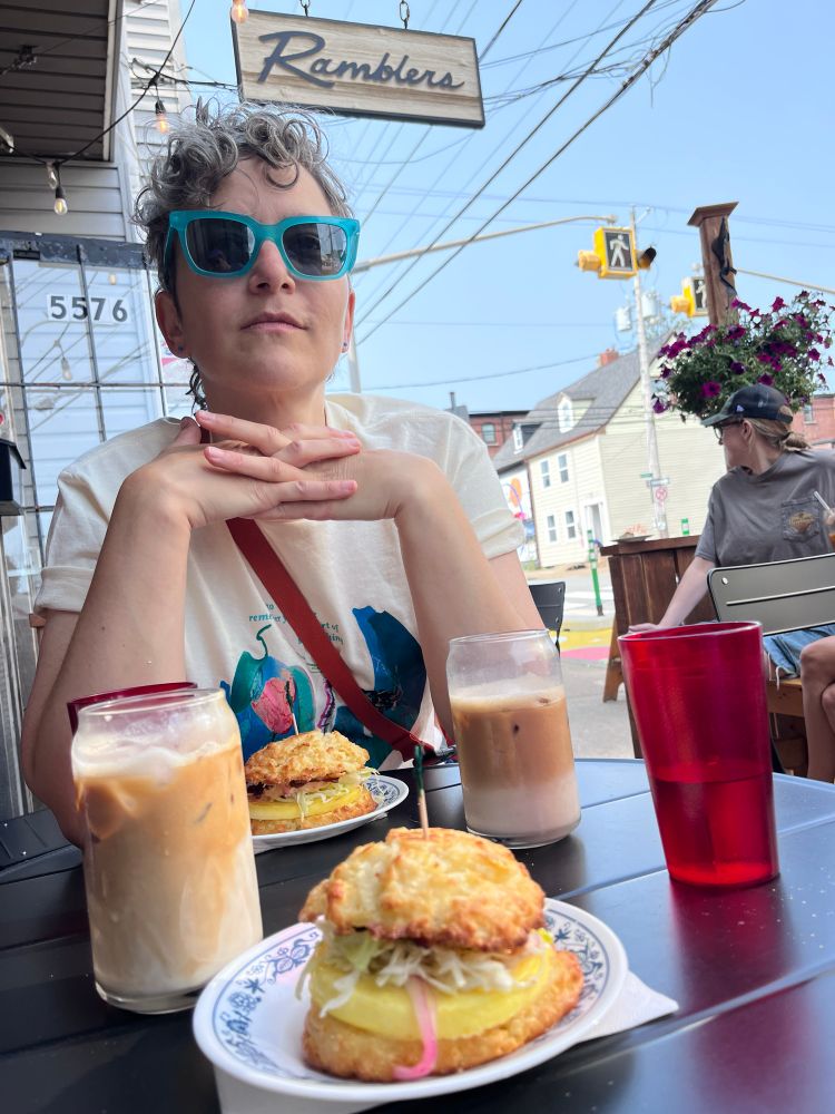 A person in blue framed sunglasses photographed from patio table level foregrounding breakfast sandwiches, iced lattes, and a red translucent tumbler of water, with the cafe sign overhead and a hanging flowering plant behind in the blue sky.