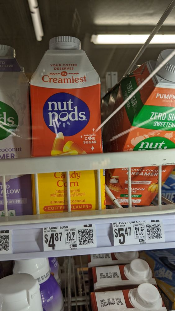 "Nut Pods" brand coffee creamer (made with Almond and Coconut Milks), Candy Corn flavored, placed on a refrigerated shelf at a Walmart, priced at $4.87; to the right, a container of "Nut Pods" brand coffee creamer (made with Almond and Coconut Milks), Pumpkin Pie flavored, priced at $5.47