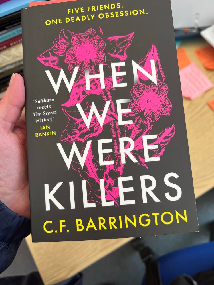 The cover of the book When We Were Killers by C.F. Barrington. There are neon pink flowers in the background of the wording and the cover is black.
