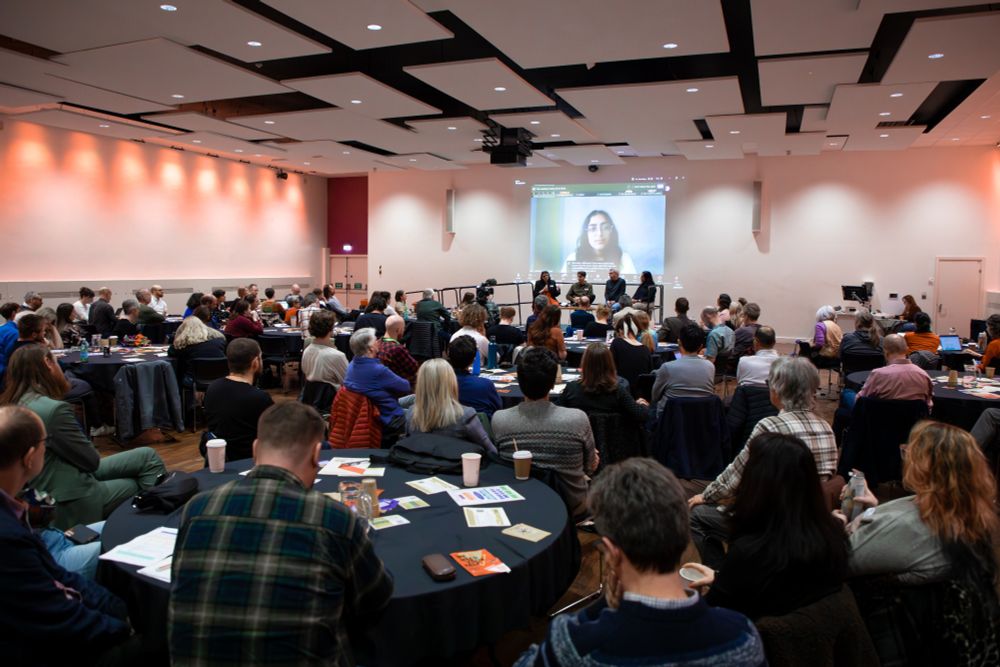 A hall full of people watching a presentation. They sit at round tables.