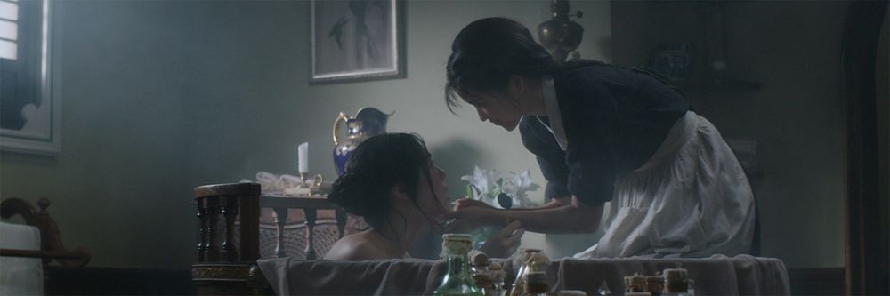 Cena do filme "A Criada", de Park Chan-wook. A imagem mostra duas mulheres interagindo em um ambiente fechado e intimista. Uma mulher está sentada em uma banheira, parcialmente submersa na água, enquanto a outra, vestida com uma blusa escura e um avental claro, se inclina sobre a banheira, segurando delicadamente o que parece ser um pequeno objeto próximo ao rosto da mulher sentada. O ambiente tem uma luz suave e difusa, com um fundo que inclui mobília de madeira, frascos e uma jarra decorativa sobre uma mesa. O tom geral da cena é sereno, com um clima de cuidado e proximidade entre as duas personagens.