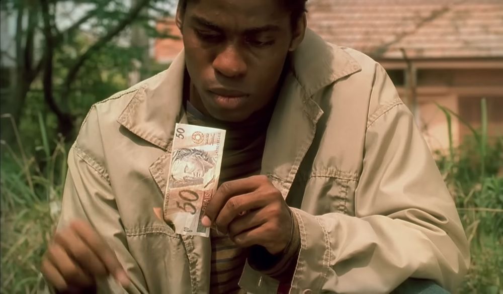 Homem jovem (Lázaro Ramos) segurando uma nota de 50 reais que começou a pegar fogo em uma mão, enquanto olha para ela com expressão séria. Ele veste um casaco bege e está ao ar livre, com uma casa desfocada e vegetação ao fundo.