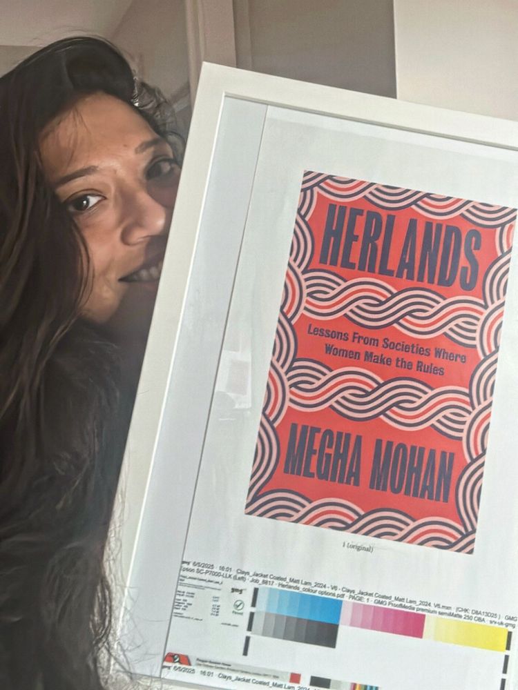 A woman with long dark hair smiles while holding a framed proof of the book “Herlands” by Megha Mohan. The book cover features bold, wavy patterns in red, white, and navy, with the subtitle “Lessons From Societies Where Women Make the Rules.”