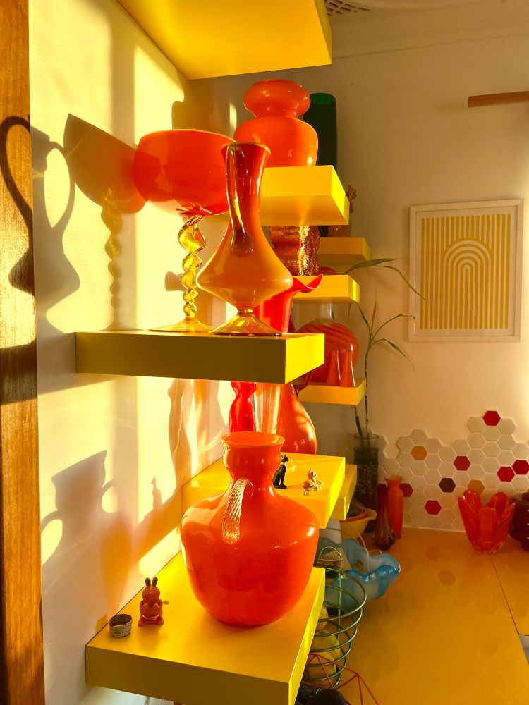 Warm yellow afternoon light hitting vintage orange glass vases displayed all on yellow shelves. The shadows onto the white wall are strong. 