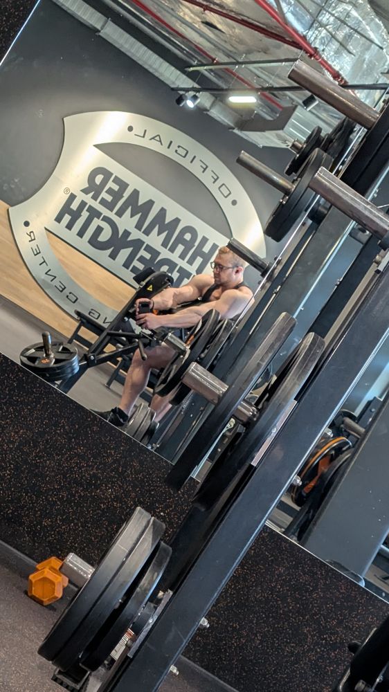 Gym mirror selfie from far away with me sitting on a preacher curl machine 