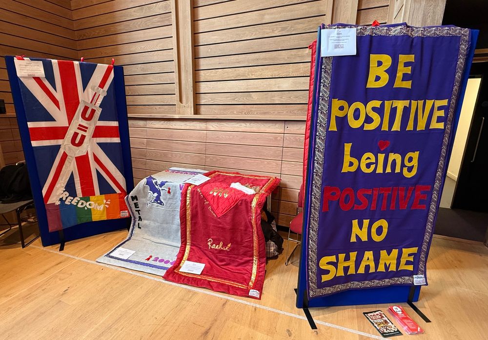 Four quilts, approximately 1 metre wide by 2 metres tall, on display in a wooden room. The one closest to the camera reads "Be positive. Being Positive, No Shame". Another has the U=U slogan on a Union Jack, with a pride flag below.