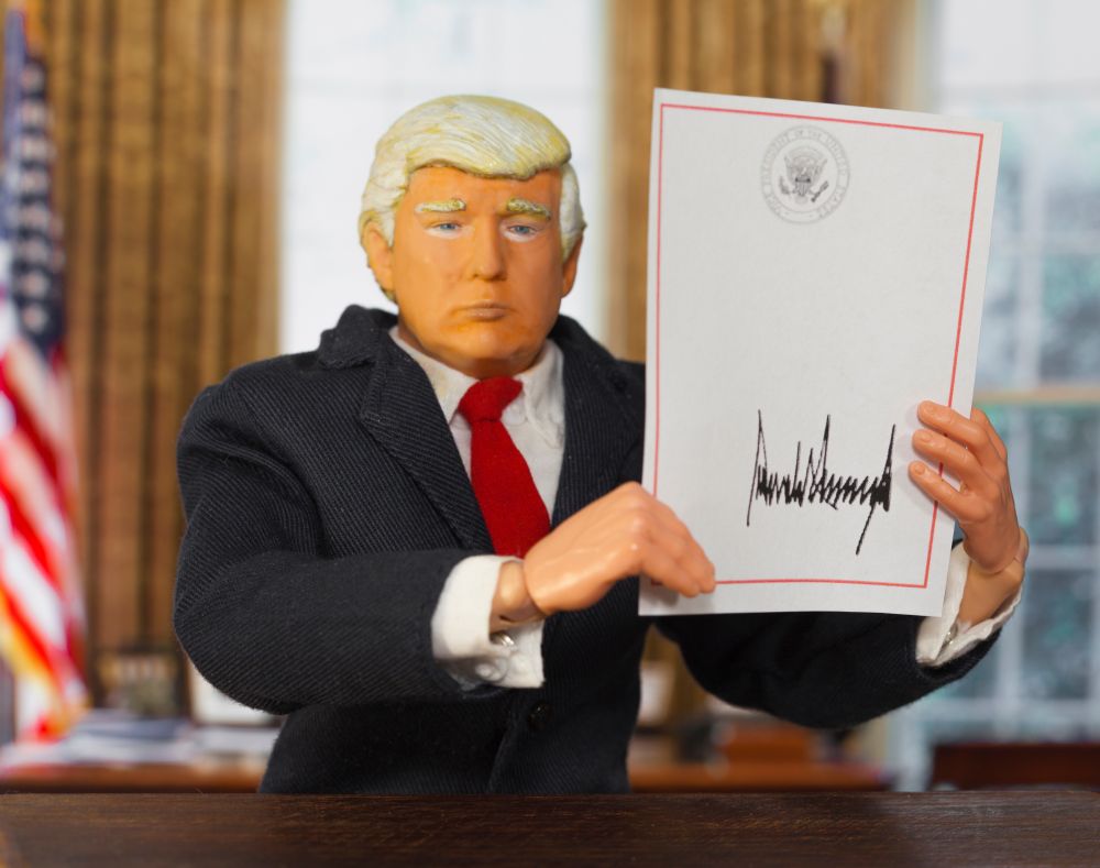 Image is copyright to Jane Doe/ Shutterstock. Shows a cartoon image of Donald Trump with a signed Executive Order