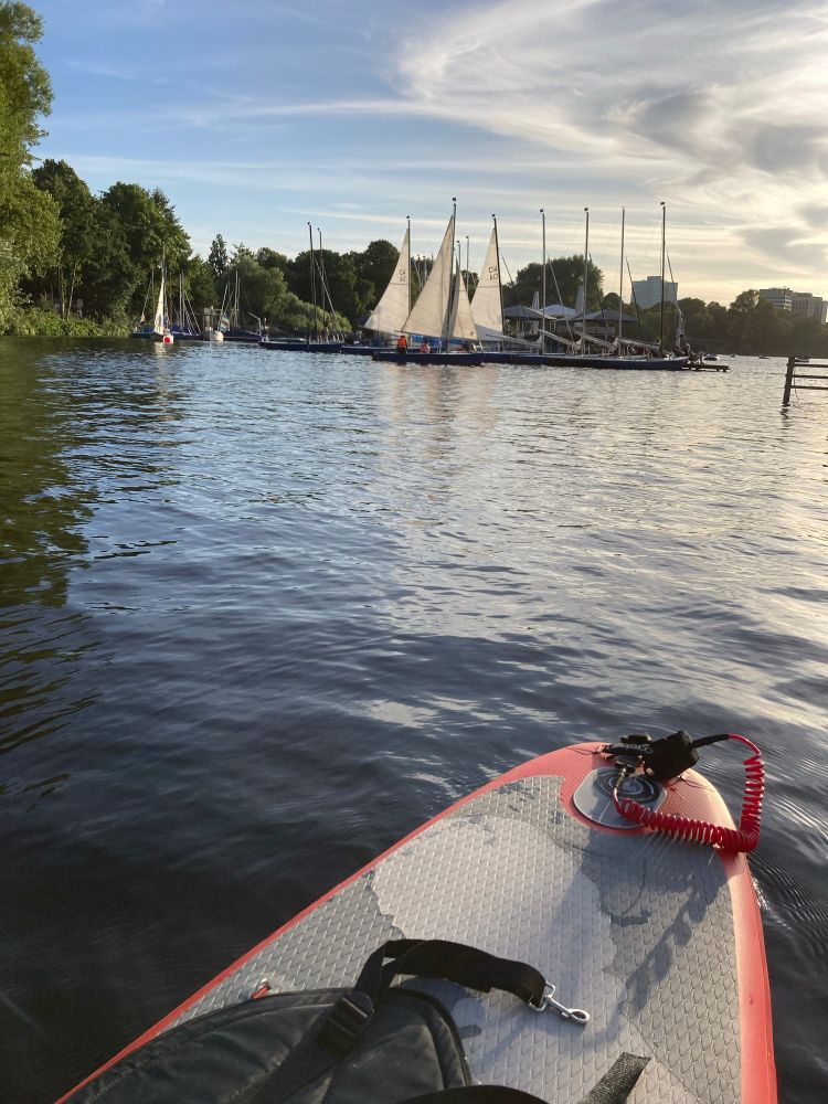 Hinteres Ende eines orangefarbenen Stand up Boards auf der Alster, etwas weiter entfernt ein Paar Segelboote am Anleger.