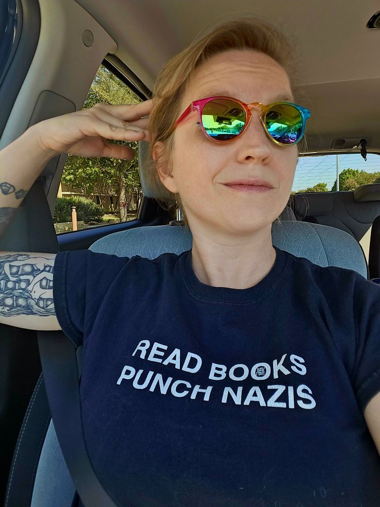 white blond lady in a car, posing with some tiny rainbow sunglasses and a black t-shirt with the words: READ BOOKS, PUNCH NAZIS