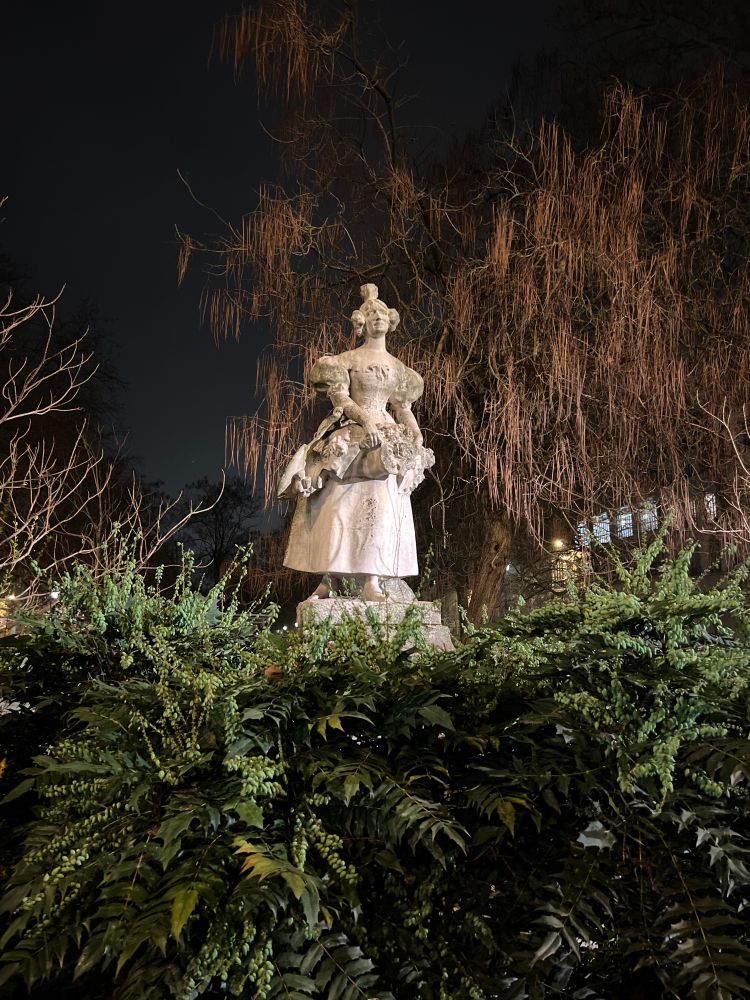 A nighttime picture of the outdoor statue called La grisette de 1830 in Paris, a young working class woman. The statue is in a park surrounded by greenery and trees.