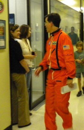 Stephen Colbert is wearing a bright orange flight suit with NASA and American flag patches walks down a hallway, holding a piece of paper. He appears to be mid-conversation or turning to speak. Two women stand by a doorway on the left, smiling, while a child and another person are visible in the background further down the hall. The setting looks like an office or lab area with framed photos on the wall.