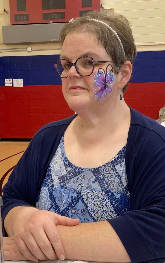A fat woman sits at a table in a gymnasium, looking at us with a mischievous side gaze.  Her arms rest on the table.  She has pixie short brown hair, adorned with a headband made of Pearl like beads.  She is wearing a top patterned in shades of blue, a navy blue cardigan, and deep purple eyeglasses.  She has a butterfly painted on her cheek in shades of purple.
