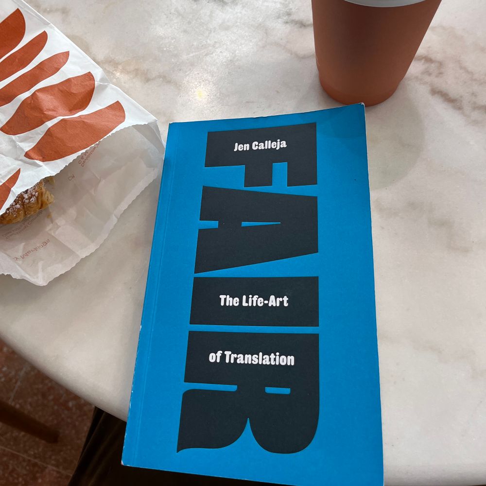 A copy of Fair: The Life-Art of Translation by Jen Calleja sits on a marble table with a coffee cup and pastry bag nearby. The cover is deep blue with the large black type of the title running vertically. 