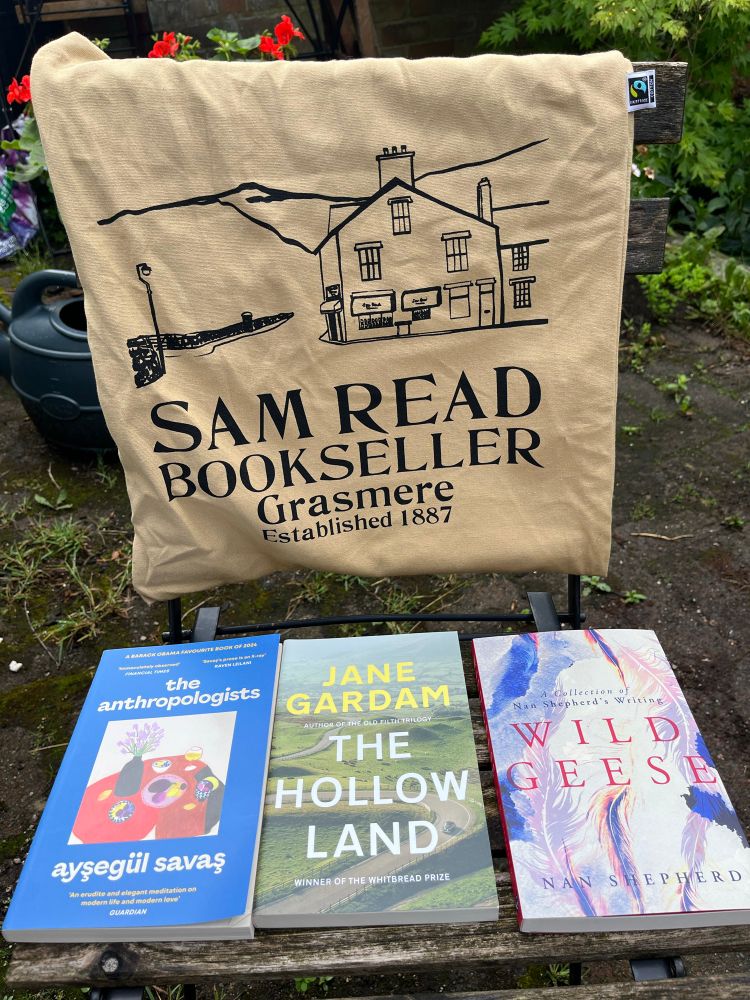 A brown tote bag hangs over the back of a chair with a black outline of a corner shop and the words Sam Read Bookseller Grasmere Established 1887. In front are three books purchased there - The Anthropologists by Ayşegül Savaş, The Hollow Land by Jane Gardam, and Wild Geese by Nan Shepherd. 