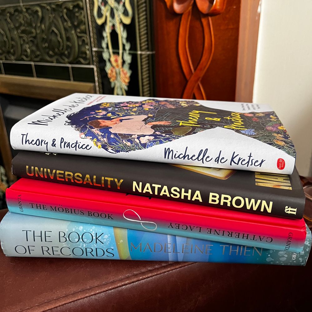 A pile of books sits on the arm of a leather chair with the spines showing: The Book of Records by Madeleine Thien, The Möbius Book by Catherine Lacey, Universality by Natasha Brown and Theory & Practice by Michelle de Kretser