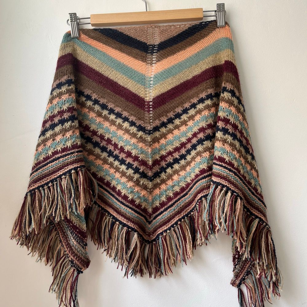 A knitted shawl on a hanger against a blank wall. The shawl has large stripes in seven different colours, then a thinner stripe sequence with a slip stitch pattern, followed by a few rows of garter stitch and ending with a tasselled fringe. 