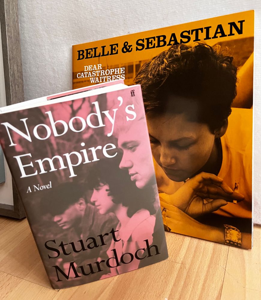 A hardcover pink copy of Nobody's Empire by Stuart Murdoch showing the side profiles of three people, against the yellow LP of Belle & Sebastian's Dear Catastrophe Waitress with its dominant photo of a young woman, eyes cast down. 