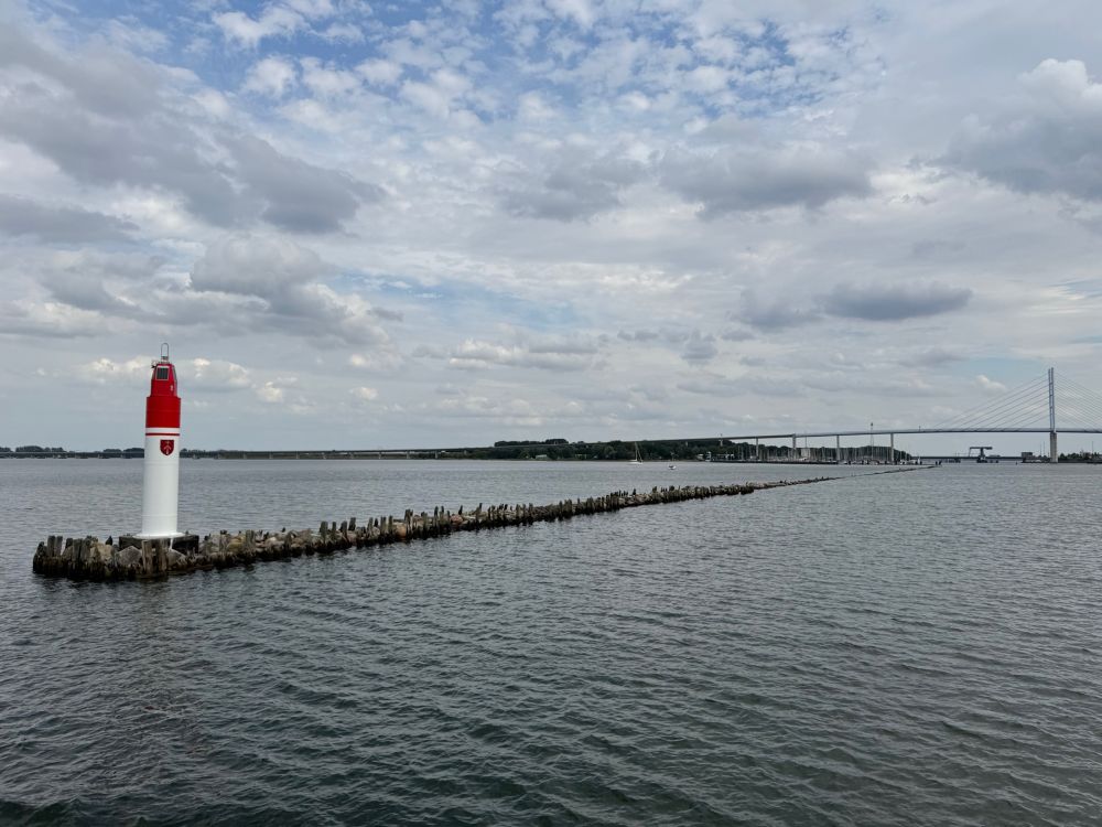 Eine Mole im Wasser, darauf ein kleines Positionslicht für die Hafeneinfahrt. Im Hintergrund überspannt eine Brücke das Wasser.