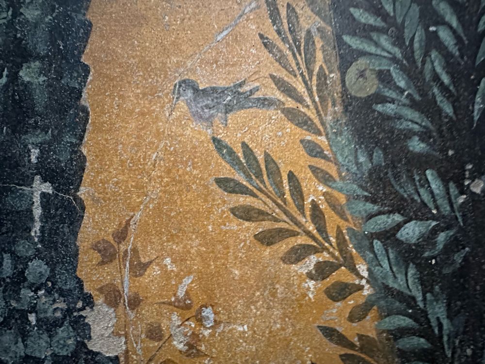 Against a yellow background a small bird teeters on a green leafed plant that sticks out of a heavier, darker green clump with a single light green fruit among its leaves