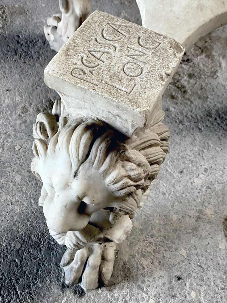 A white marble table leg in the form of a lion head and descending into a claw as the foot. The view is from above and since there is no table top the inscription of P. Casca Long can be seen engraved into the table top support.