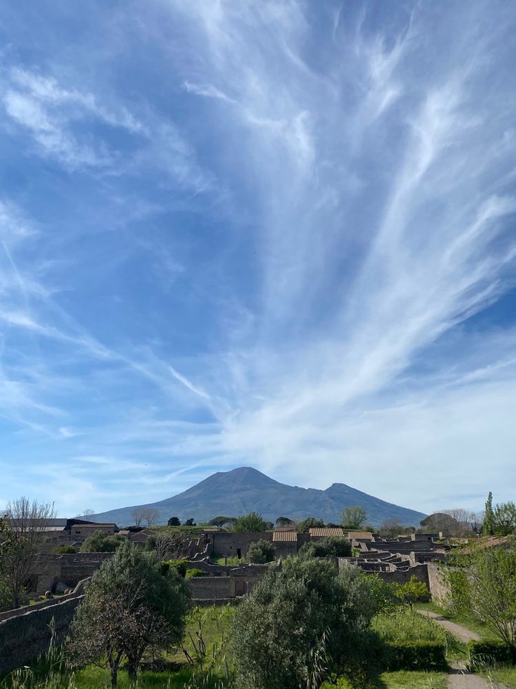 View of the outline of Vesuvius looming above the houses of ancient Pompeii and above clouds swirl against a blue sky seemingly like an explosion from the cone of the volcano