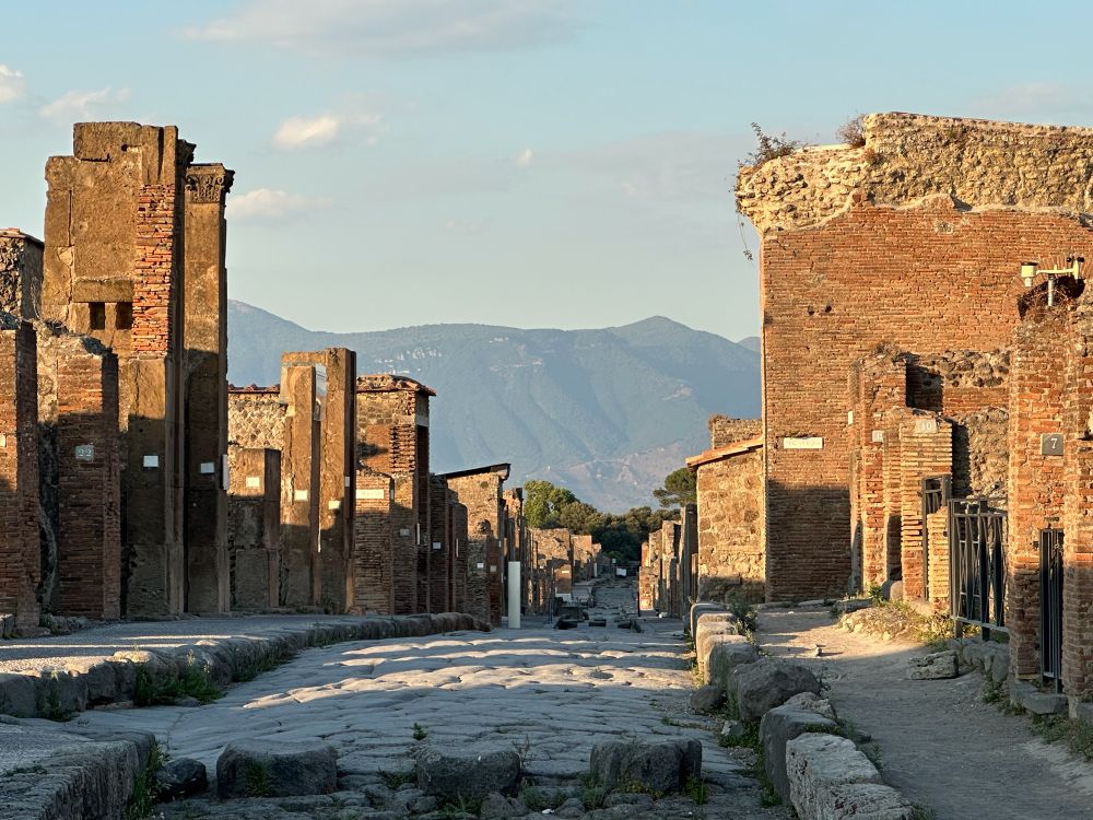 View down an empty street with warm evening sunshine illuminating the walls. Mountains form the backdrop with a soft blue sky