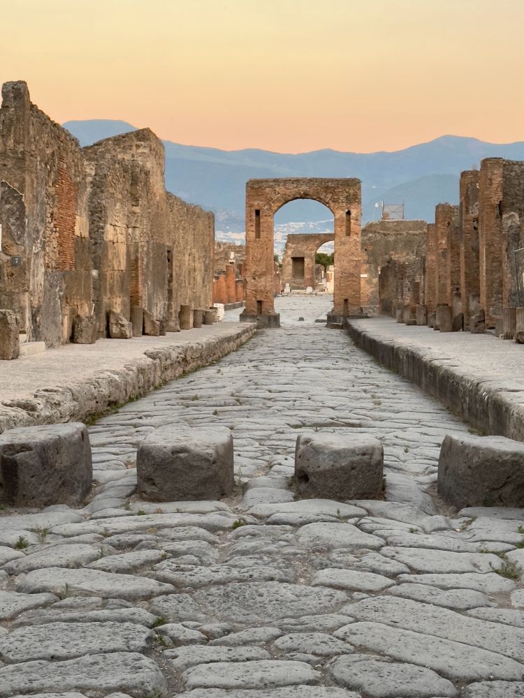 View down the via di Mercurio towards the arch of Caligula and the forum beyond. The sky above the Lattari hills glows a soft hazy yellowy orangey