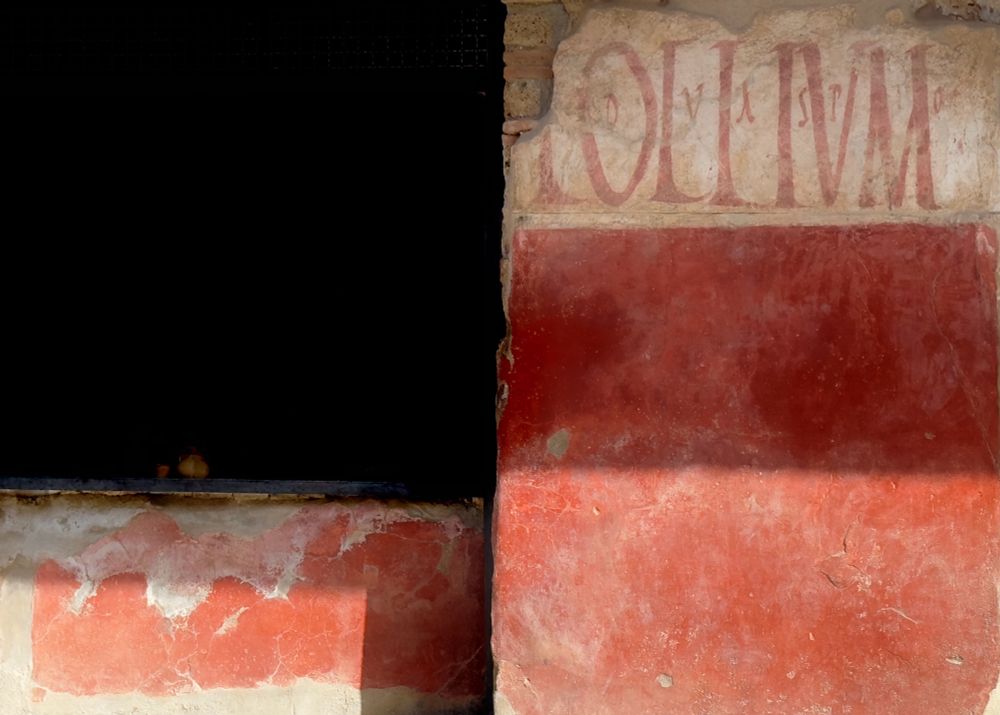 An ancient bar frontage decorated in red plaster. The bar counter on the left is slightly cast in rectangular shadow and the depths of the bar remain shrouded in darkness. The facade on the right of the image has an upper portion in white with red lettering and the shadow falling across it all aligns with the height of the bar counter. A geometric play of light, dark and shadows.