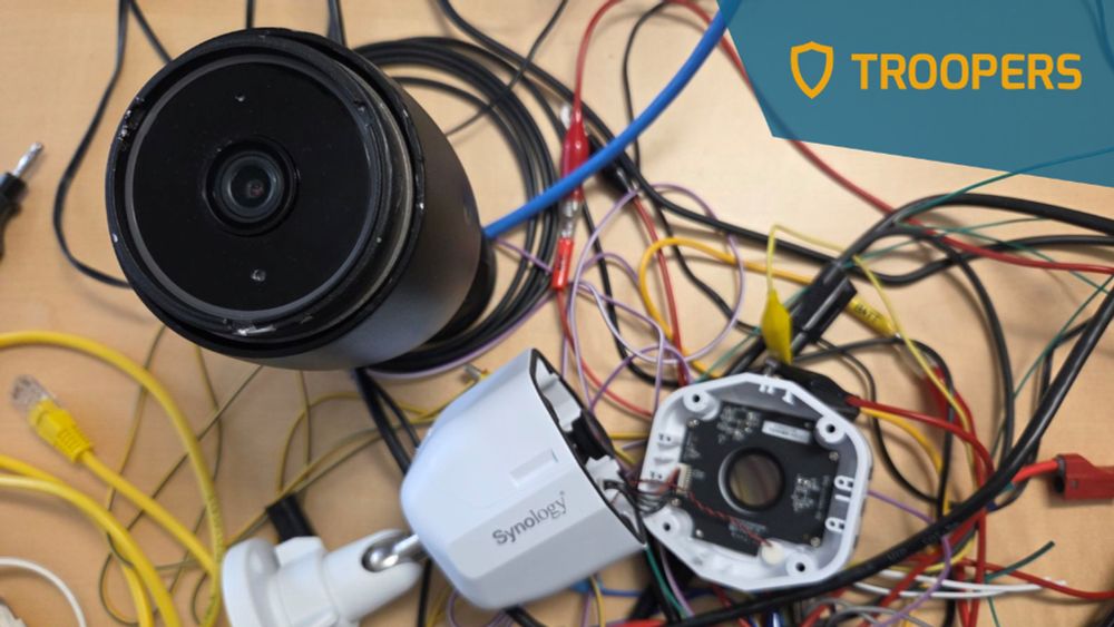 High-resolution photo of Compass Security’s IoT and industrial penetration-testing workspace: on a light wooden workbench a large-lens, black surveillance camera sits half-disassembled beside its white Synology® housing, revealing the internal printed-circuit board, image sensor and ribbon connectors targeted during firmware extraction and vulnerability analysis. A chaotic web of multicolored diagnostic leads, Ethernet patch cables, alligator clips, UART/serial breakout wires and power adapters snakes across the table, illustrating real-world hardware hacking, fault-injection and secure-boot bypass techniques used in red-team assessments of networked CCTV, smart-factory and critical OT devices. The blue pentagonal TROOPERS25 shield logo occupies the upper-right corner, signalling that this lab scene supports Compass Security’s conference presentation on Pwn2Own-grade research into surveillance-camera exploits, remote-code-execution vectors and zero-day discovery. The image underscores expert penetration-testing methodology—threat modeling, reverse engineering, embedded Linux analysis, secure-element probing and API fuzzing.