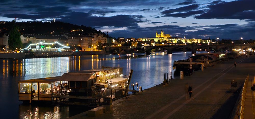 Prague by night along the Vltava river. Boats decorated with string lights line the river, and the Prague castle is all lit up in the background.