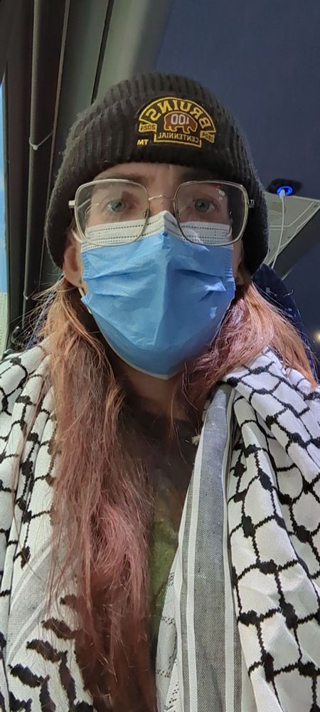Woman in glasses, a blue mask, and a Bruins beanie. She's wearing a keffiyeh around her neck.