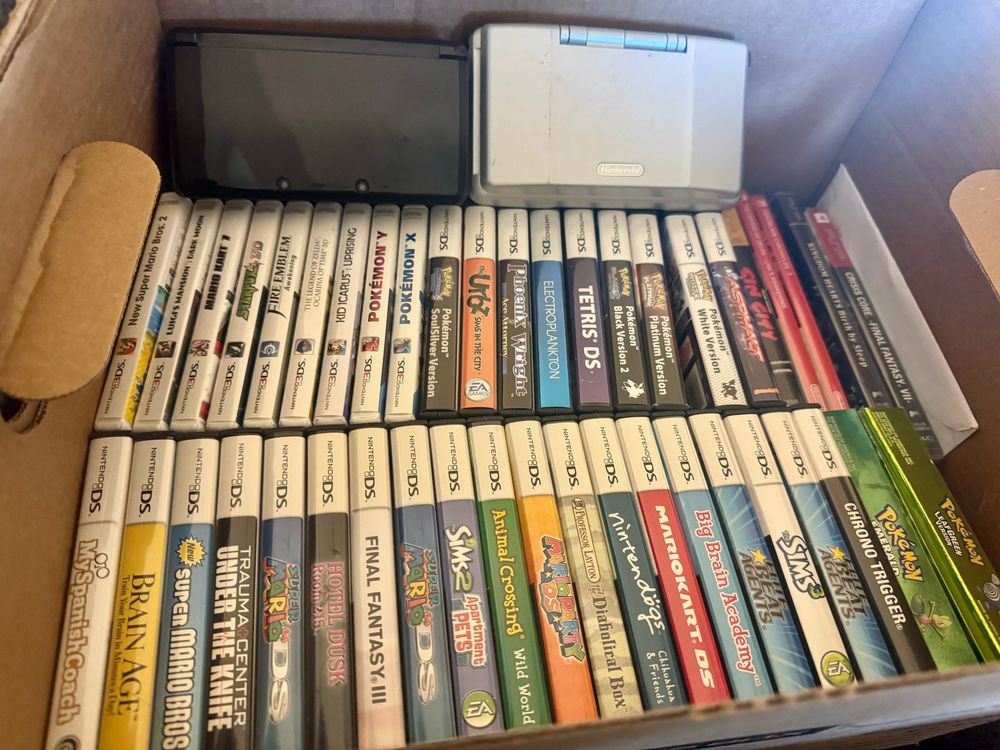 The image shows a cardboard box filled with various Nintendo DS game cartridges and a handheld console. At the top left, a Nintendo DS device is visible, partially obscured. Below it, a neatly organized row of game cartridges features titles such as "New Super Mario Bros," "Fire Emblem," "Pokémon Y," "Tetris DS," and "Final Fantasy III." The cartridges are arranged with their labels facing outward, showcasing a variety of popular games across different genres, including adventure, puzzle, and role-playing games. The box also contains additional titles like "Animal Crossing," "Mario Kart DS," and "Chrono Trigger." The overall scene suggests a collection belonging to a gaming enthusiast, highlighting the nostalgia and variety of the Nintendo DS library. The box appears to be in a well-lit environment, emphasizing the colorful artwork on the game cases.