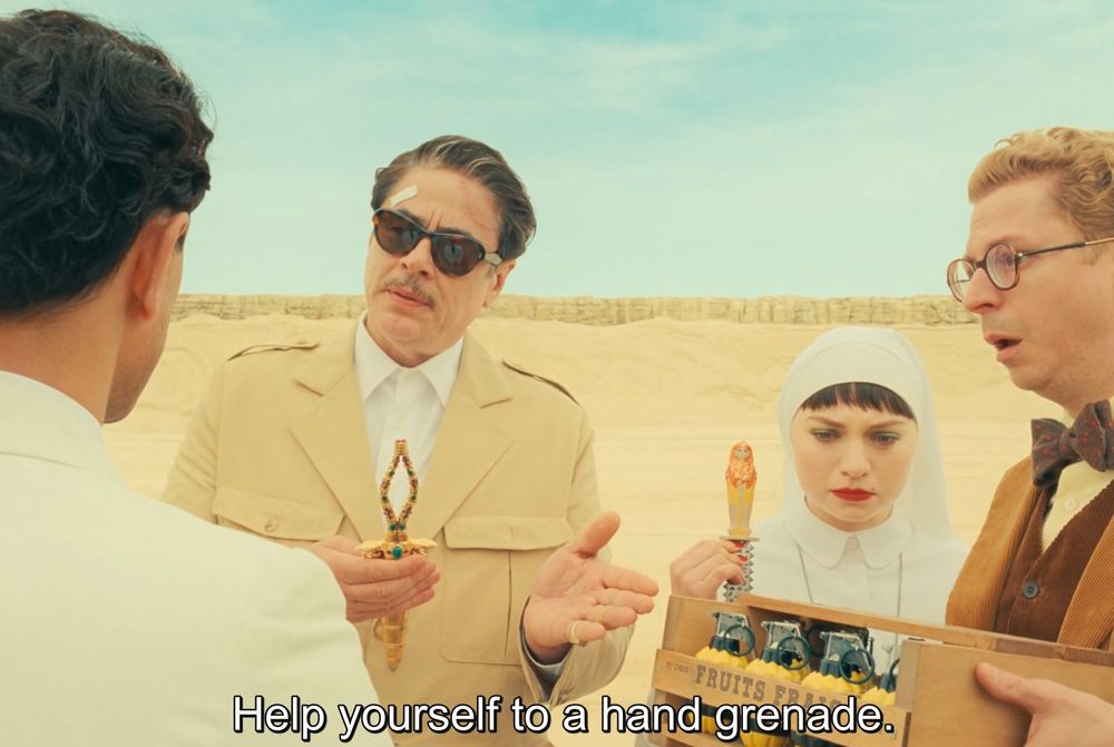 A still from The Phoenician Scheme, standing in the desert with his daughter who’s a nun, and his administrative secretary, Benicio Del Toro as businessman Zsa Zsa Kordo, says, “Help yourself to a hand grenade.”