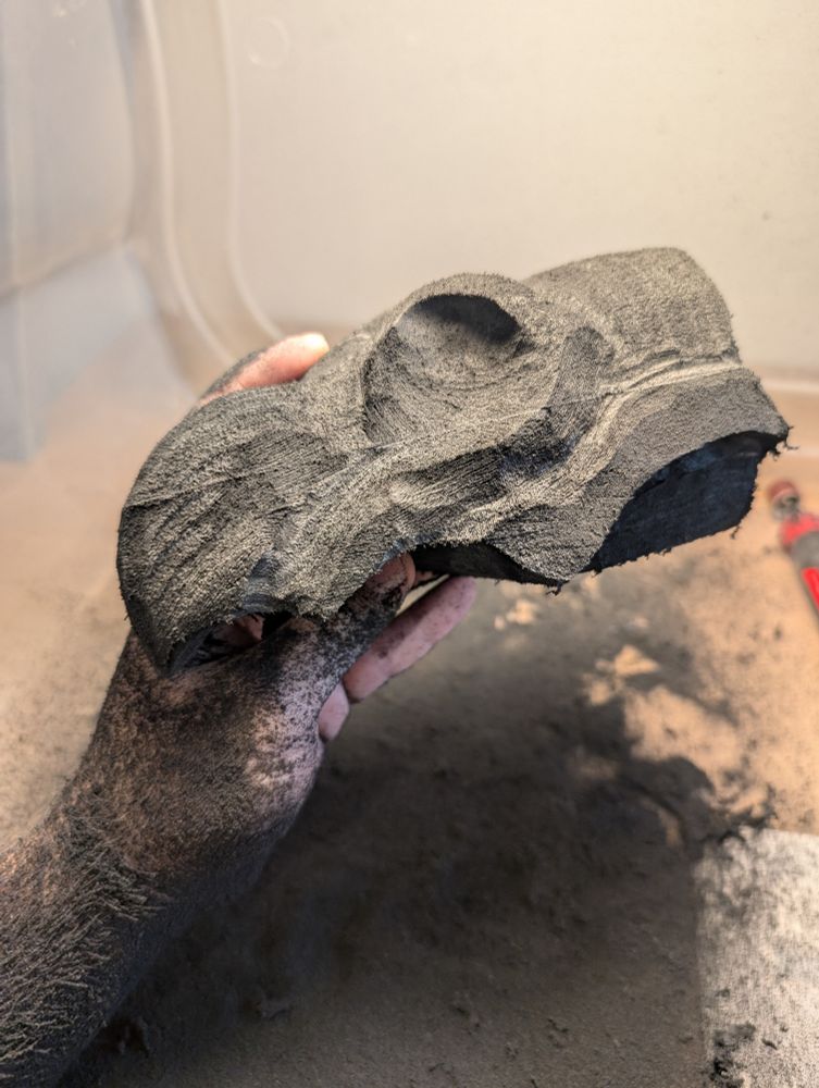The same hand, now covered in a dark dust, holds the same lump of foam, now a lot more carved into. It looks kind of like a skull with a beak at this point
