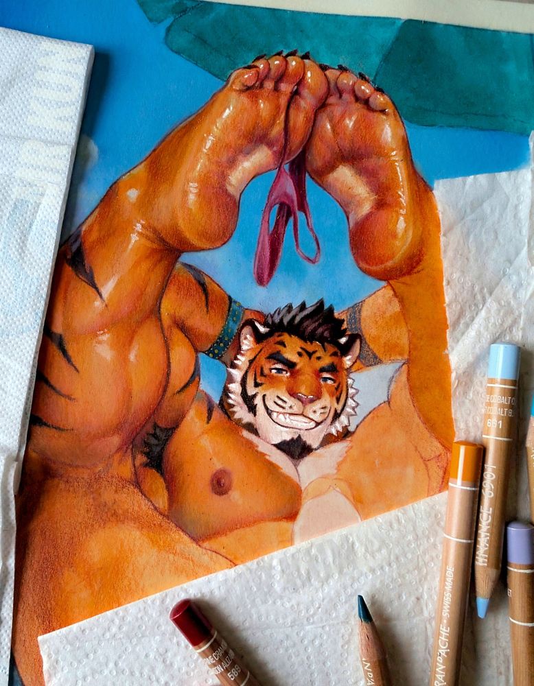 A muscular anthropomorphic tiger lying on his back with his large feet raised up, holding a tiny pink thong between his toes. His fur is orange with black stripes, and he wears a confident, teasing smile. The artwork is traditional, surrounded by colored pencils and tissues, showing it's still in progress.