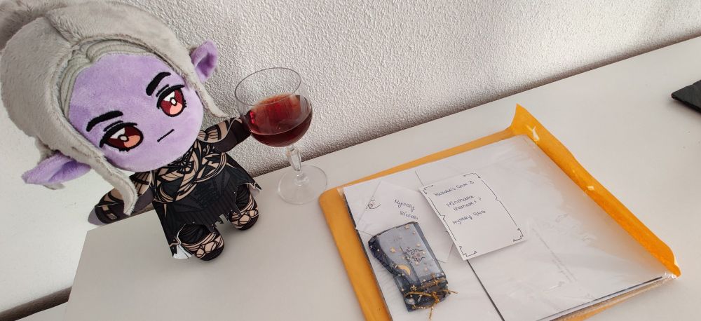 A Minthara Baenre plushie inspecting a mystery bag of Baldur's Gate 3 fan merch, next to her a glass of wine.