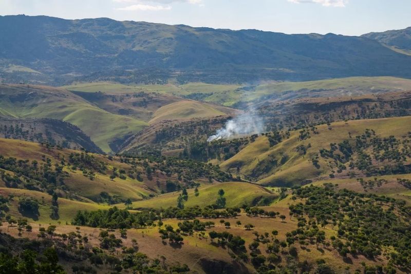 Panoramic view of Madagascar’s Itremo Massif showing rolling hills, patchy forest cover, and a plume of smoke rising from the valley floor.