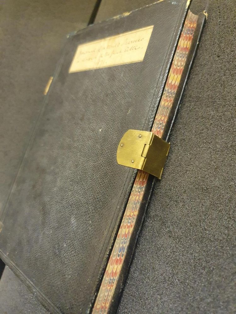  A side view of a bound volume with a metal clasp. The volume has a label affixed to its cover and the writing on it is blurry. The page edges are marbled. 