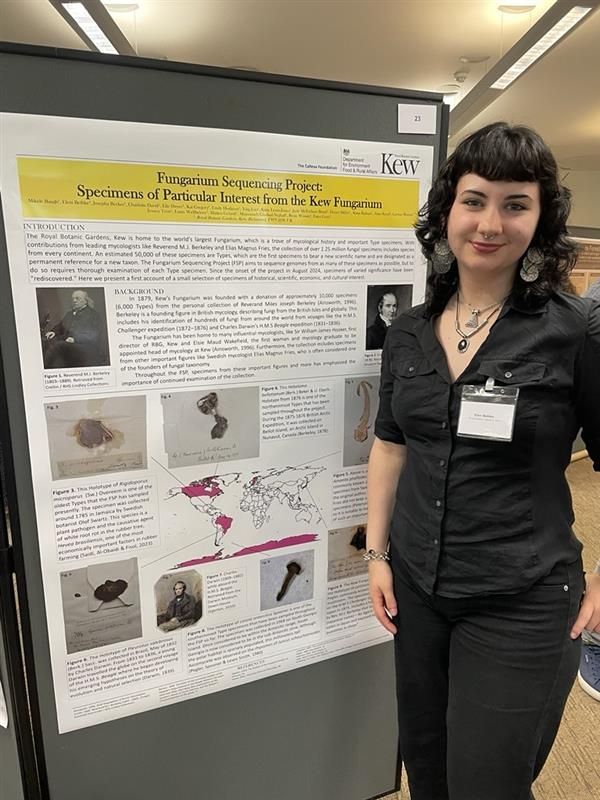 A researcher stands next to a poster titled “Fungarium Sequencing Project: Specimens of Particular Interest from the Kew Fungarium”, featuring specimen images, a world map, and historical figures including Darwin.
