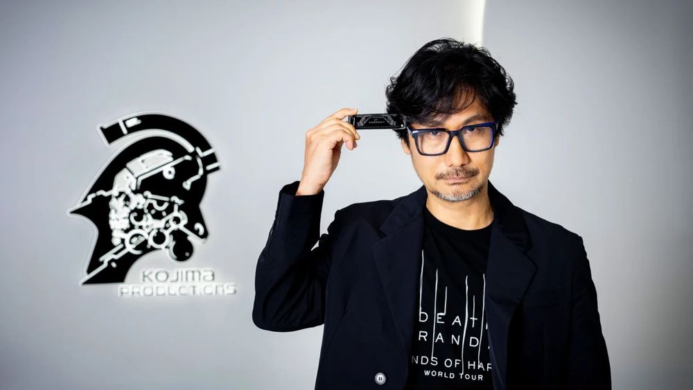 Kojima in front of a Kojima productions logo with a harmonica, t-shirt and sports coat combo