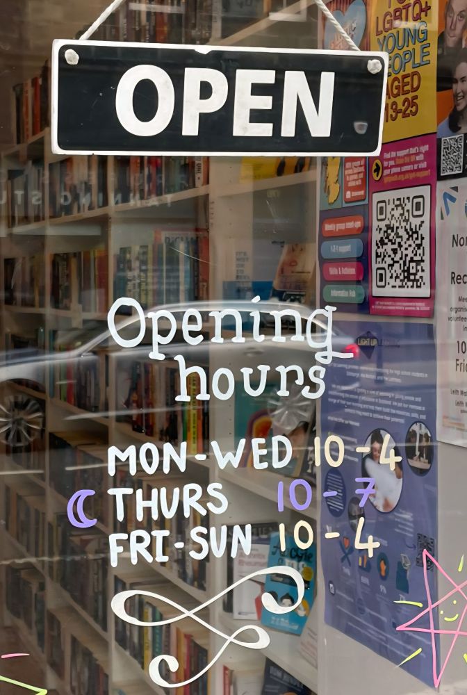 Photograph of the front window of a bookshop, in which hangs an OPEN sign, with the opening hours of Monday to Wednesday 10 to 4, Thursday 10 to 7 and Friday to Sunday 10 to 4. 