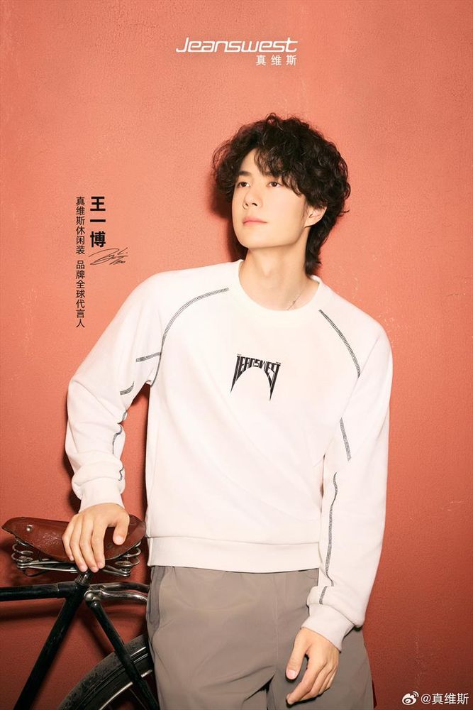 Wang Yibo in a cream windcheater and brown trousers, leans up on a push bike, with a red ochre wall behind him.