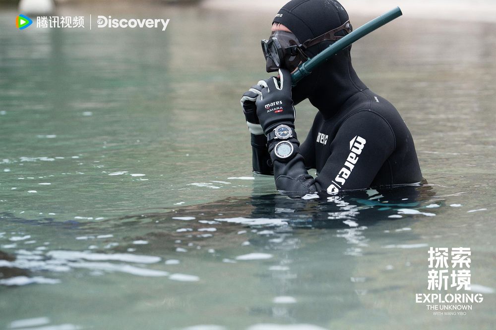 Wang Yibo in a black wetsuit adjusting his snorkel, chest deep in water, seen from the side.
