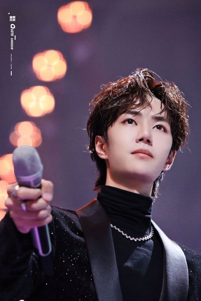 Wang Yibo in a black jacket over a black turtle neck with a silver neck chain, viewed from below, holding out a microphone.