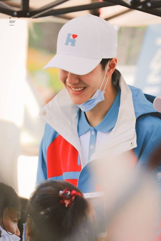 Wang Yibo in white blue and red top, white cap obscuring his eyes, blue medical face mask pulled down under his chin, smiling.