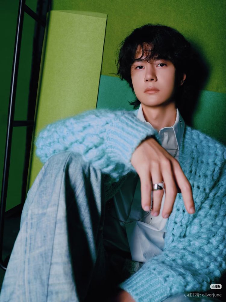Wang Yibo with curly hair, a super soft blue cardigan and grey flannel trousers, sitting up against a green wall.