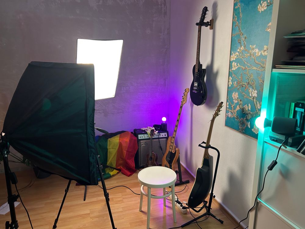 A “behind the scenes shot of an improvised stage for a video shot filled with soft boxes and other lights, guitars are visible in the background while a chair is standing in the middle of the scene
